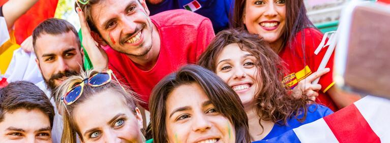 Football supporter taking a selfie at stadium. Friends cheering and watching soccer match together at stadium. International and multiracial group with multicolored t-shirts having fun.